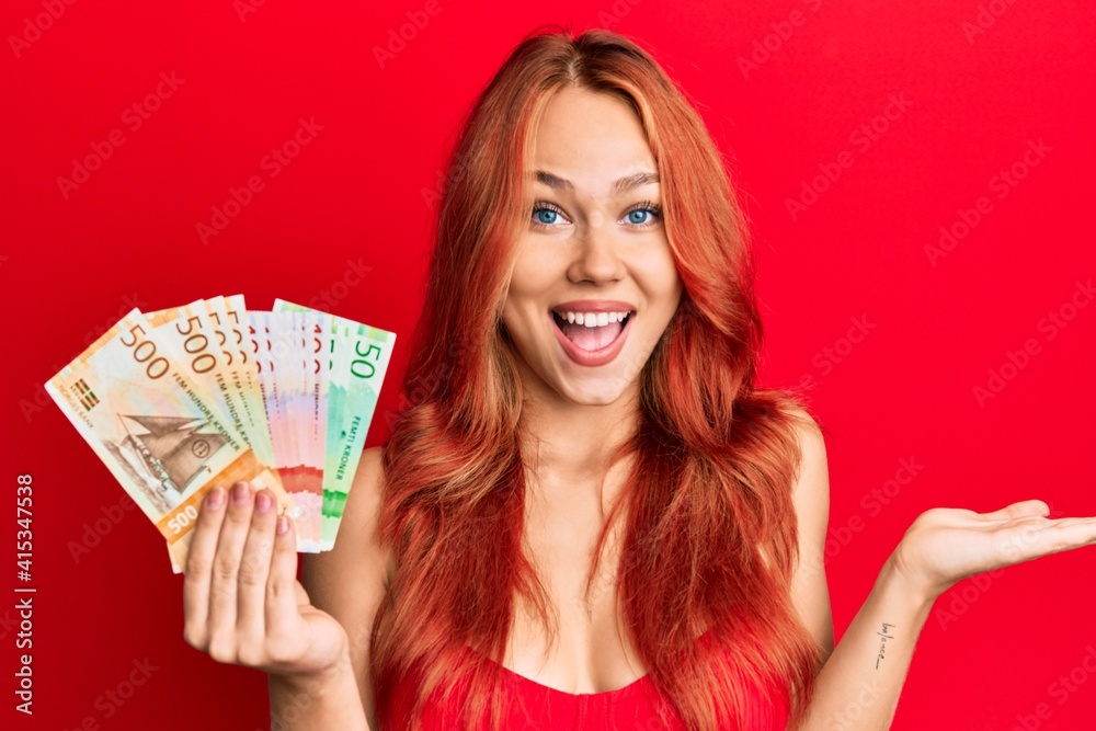 Young beautiful redhead woman holding norwegian krone banknotes celebrating victory with happy smile and winner expression with raised hands Young beautiful redhead woman holding norwegian krone banknotes celebrating victory with happy smile and winner expression with raised hands