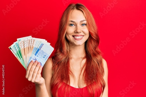 Young beautiful redhead woman holding south korean won banknotes looking positive and happy standing and smiling with a confident smile showing teeth