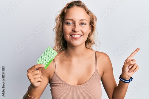 Beautiful caucasian woman holding birth control pills smiling happy pointing with hand and finger to the side