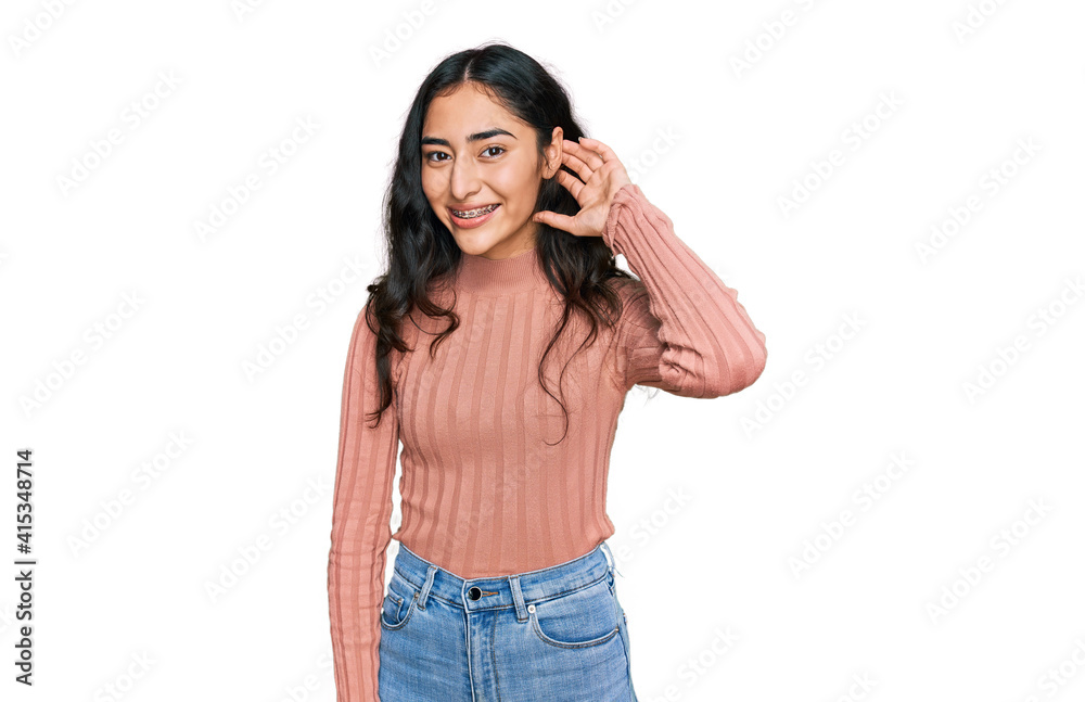 Hispanic teenager girl with dental braces wearing casual clothes smiling with hand over ear listening an hearing to rumor or gossip. deafness concept. Hispanic teenager girl with dental braces wearing casual clothes smiling with hand over ear listening an hearing to rumor or gossip. deafness concept.