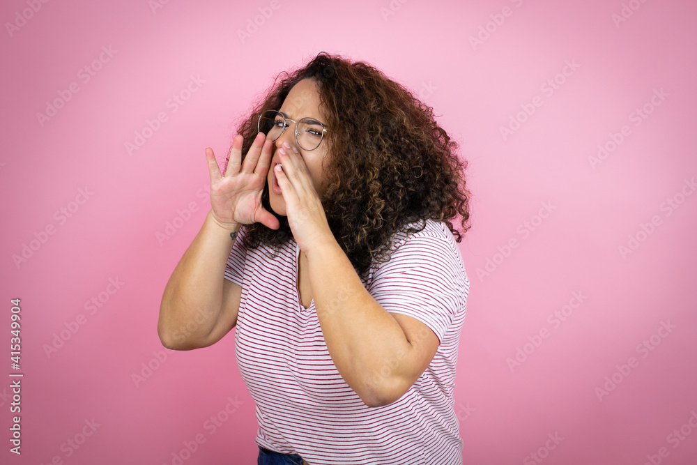 Young african american woman wearing red stripes t-shirt over pink background shouting and screaming loud to side with hands on mouth Young african american woman wearing red stripes t-shirt over pink background shouting and screaming loud to side with hands on mouth