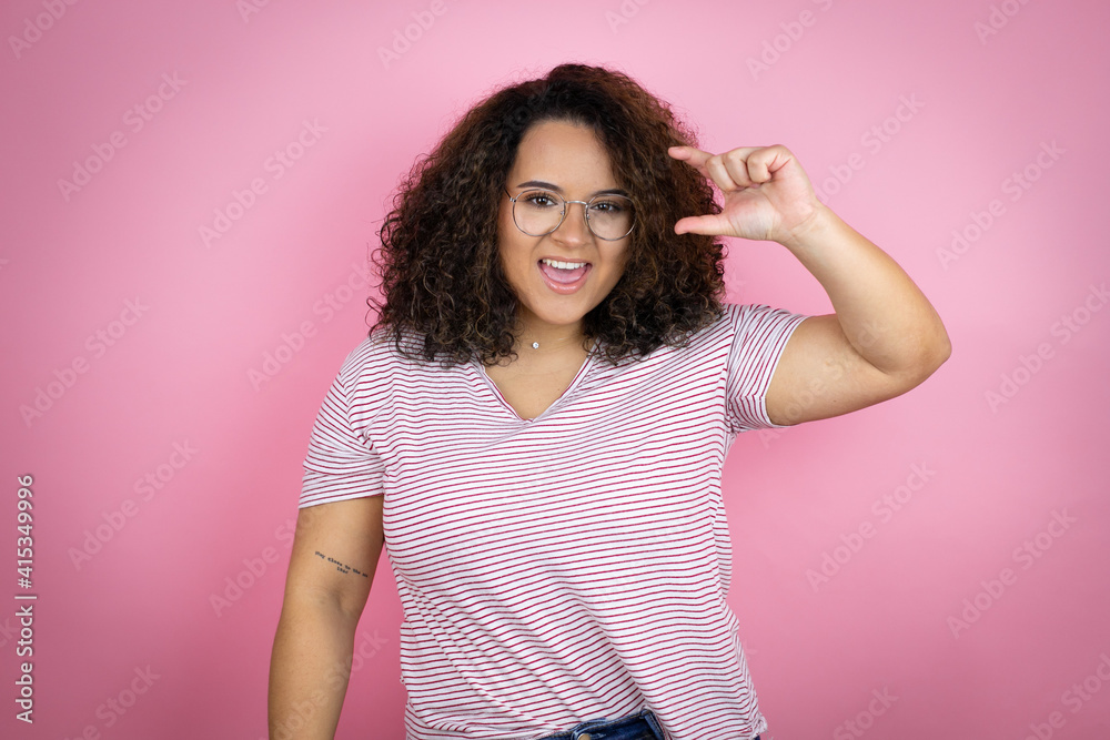 Young african american woman wearing red stripes t-shirt over pink background smiling and confident gesturing with hand doing small size sign with fingers . Measure concept. Young african american woman wearing red stripes t-shirt over pink background smiling and confident gesturing with hand doing small size sign with fingers . Measure concept.