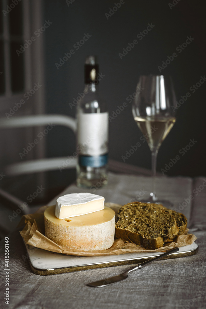 A glass of white wine, a bottle, several kinds of cheese and bread on a cutting board. Vertical lifestyle photo