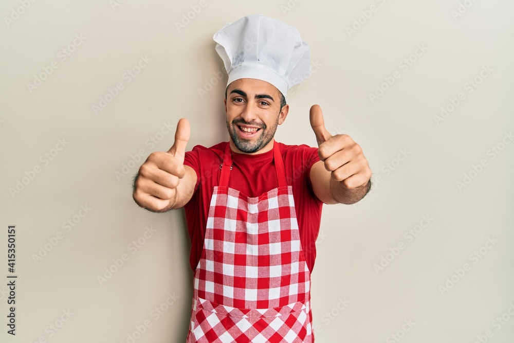 Young hispanic man wearing baker uniform approving doing positive gesture with hand, thumbs up smiling and happy for success. winner gesture. Young hispanic man wearing baker uniform approving doing positive gesture with hand, thumbs up smiling and happy for success. winner gesture.