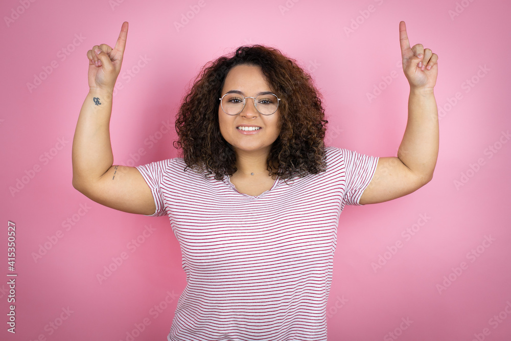 Young african american woman wearing red stripes t-shirt over pink background smiling, looking at the camera and pointing up with fingers and raised arms Young african american woman wearing red stripes t-shirt over pink background smiling, looking at the camera and pointing up with fingers and raised arms