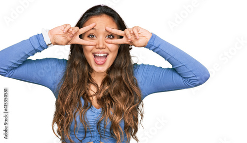 Young hispanic girl wearing casual clothes doing peace symbol with fingers over face, smiling cheerful showing victory