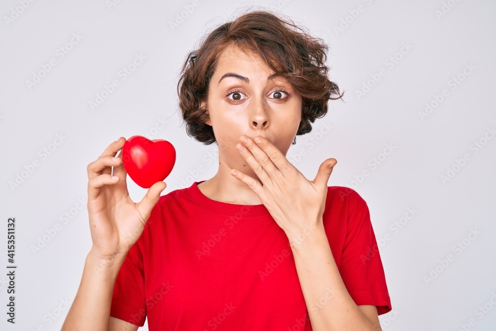 Young hispanic woman holding heart covering mouth with hand, shocked and afraid for mistake. surprised expression Young hispanic woman holding heart covering mouth with hand, shocked and afraid for mistake. surprised expression