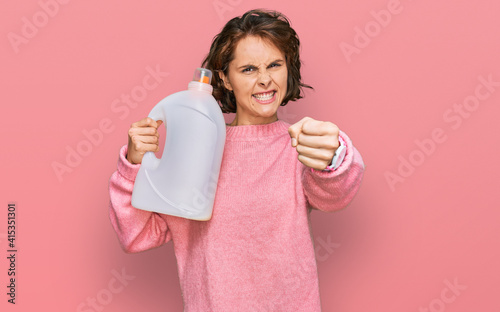 Young hispanic woman doing laundry holding detergent bottle annoyed and frustrated shouting with anger, yelling crazy with anger and hand raised