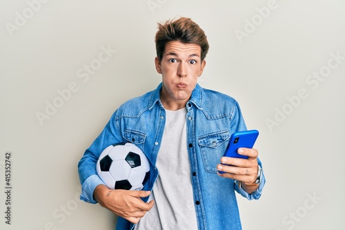 Handsome caucasian man holding football ball looking at smartphone puffing cheeks with funny face. mouth inflated with air, catching air.