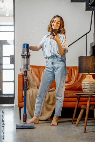 Billede på lærred Happy woman with cordless vacuum cleaning floor and using phone at home in stylish interior design of living room
