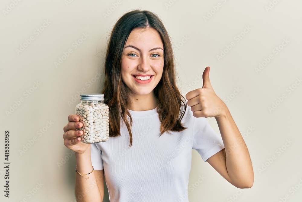Young brunette woman holding bottle with raw white beans smiling happy and positive, thumb up doing excellent and approval sign Young brunette woman holding bottle with raw white beans smiling happy and positive, thumb up doing excellent and approval sign