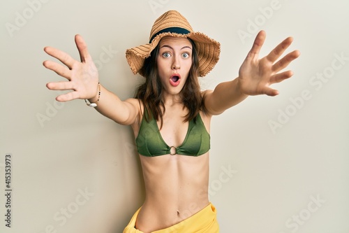 Young brunette woman wearing bikini and hat with open arms hugging afraid and shocked with surprise and amazed expression, fear and excited face.