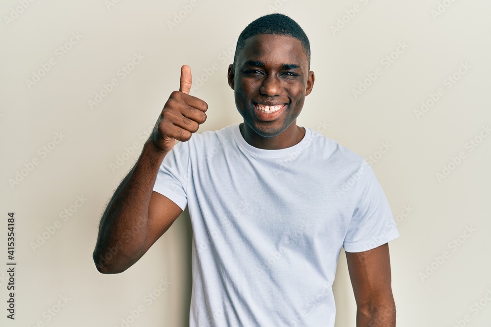 Young african american man wearing casual white t shirt smiling happy and positive, thumb up doing excellent and approval sign Young african american man wearing casual white t shirt smiling happy and positive, thumb up doing excellent and approval sign