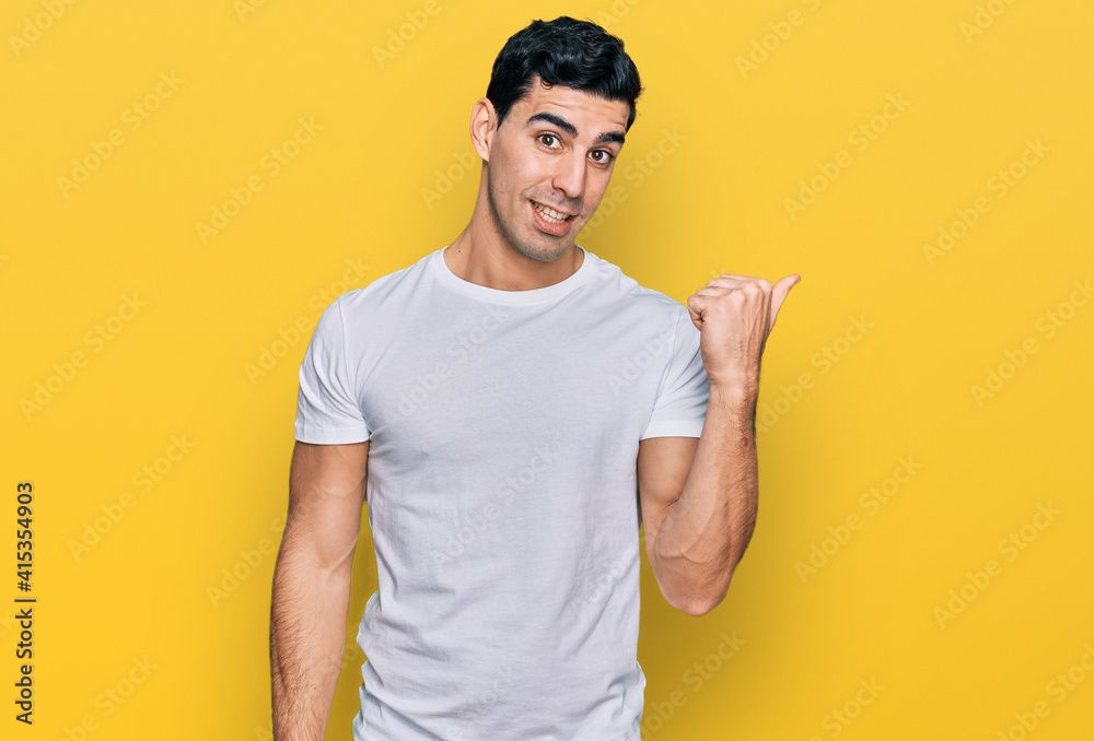 Handsome hispanic man wearing casual white t shirt smiling with happy face looking and pointing to the side with thumb up. Handsome hispanic man wearing casual white t shirt smiling with happy face looking and pointing to the side with thumb up.