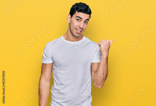 Handsome hispanic man wearing casual white t shirt smiling with happy face looking and pointing to the side with thumb up.