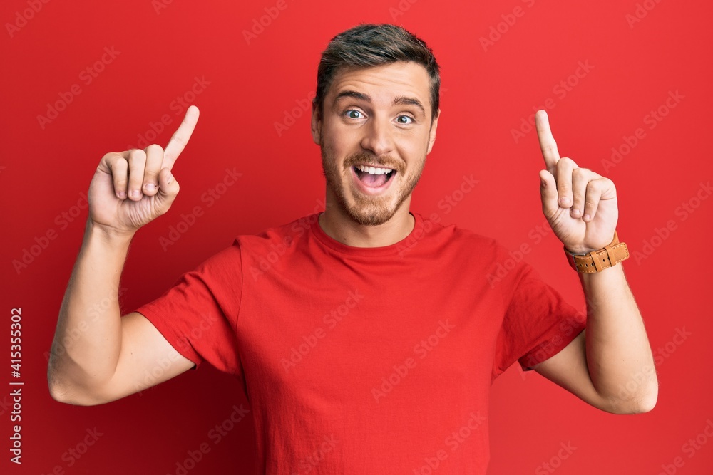 Handsome caucasian man wearing casual red tshirt smiling amazed and surprised and pointing up with fingers and raised arms. Handsome caucasian man wearing casual red tshirt smiling amazed and surprised and pointing up with fingers and raised arms.