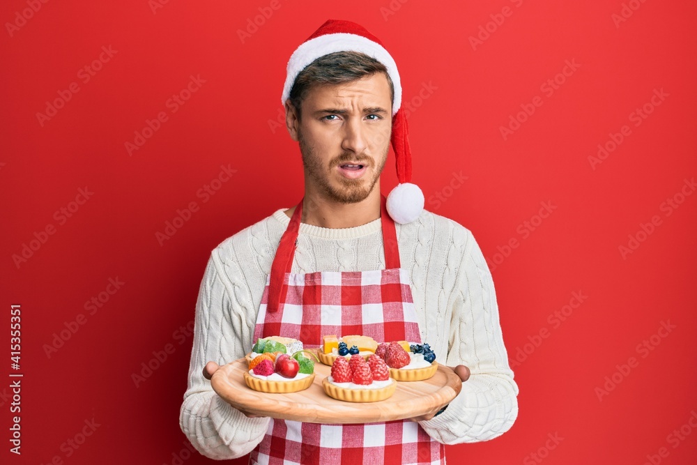 Handsome caucasian man wearing baker apron and christmas hat holding cakes in shock face, looking skeptical and sarcastic, surprised with open mouth Handsome caucasian man wearing baker apron and christmas hat holding cakes in shock face, looking skeptical and sarcastic, surprised with open mouth