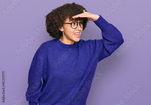 Young hispanic girl wearing casual winter sweater and glasses very happy and smiling looking far away with hand over head. searching concept. © Krakenimages.com