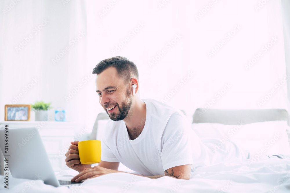 Handsome young modern bearded man works on the laptop while lying on the big white bed at home in the morning Handsome young modern bearded man works on the laptop while lying on the big white bed at home in the morning