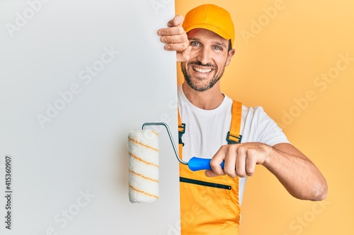 Fototapeta Naklejka Na Ścianę i Meble -  Young handsome man holding roller painter over white banner smiling with a happy and cool smile on face. showing teeth.
