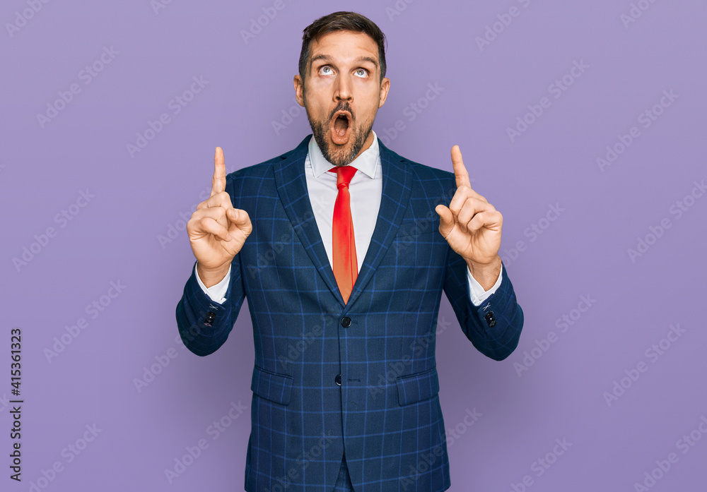 Handsome man with beard wearing business suit and tie amazed and surprised looking up and pointing with fingers and raised arms. Handsome man with beard wearing business suit and tie amazed and surprised looking up and pointing with fingers and raised arms.
