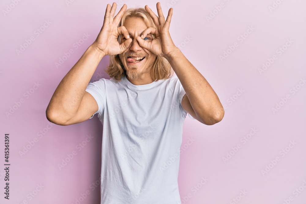 Caucasian young man with long hair wearing casual white t shirt doing ok gesture like binoculars sticking tongue out, eyes looking through fingers. crazy expression. Caucasian young man with long hair wearing casual white t shirt doing ok gesture like binoculars sticking tongue out, eyes looking through fingers. crazy expression.