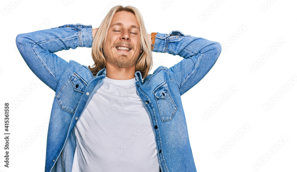 Caucasian man with blond long hair wearing casual denim jacket relaxing and stretching, arms and hands behind head and neck smiling happy Caucasian man with blond long hair wearing casual denim jacket relaxing and stretching, arms and hands behind head and neck smiling happy