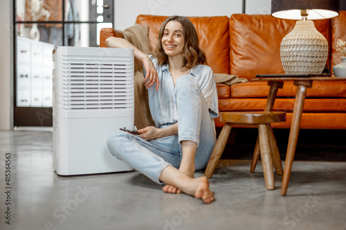 Pretty woman sitting near air purifier and moisturizer appliance near sofa monitoring air quality in phone. Health microclimate at home concept. Looking at camera.