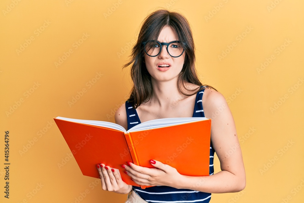 Young beautiful caucasian girl reading a book wearing glasses clueless and confused expression. doubt concept. Young beautiful caucasian girl reading a book wearing glasses clueless and confused expression. doubt concept.