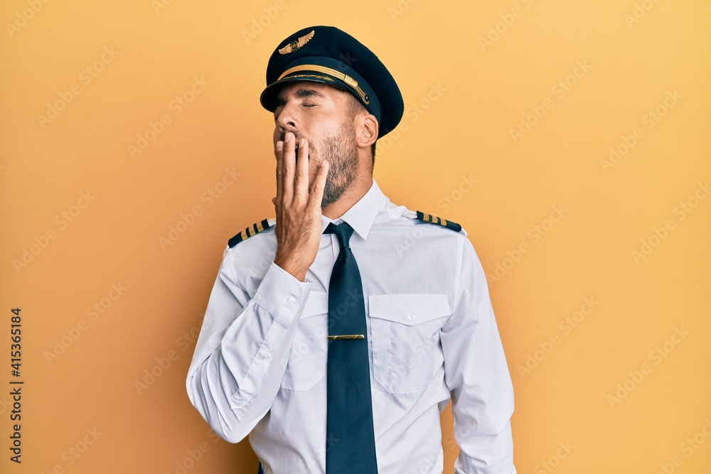 Handsome hispanic man wearing airplane pilot uniform bored yawning tired covering mouth with hand. restless and sleepiness.