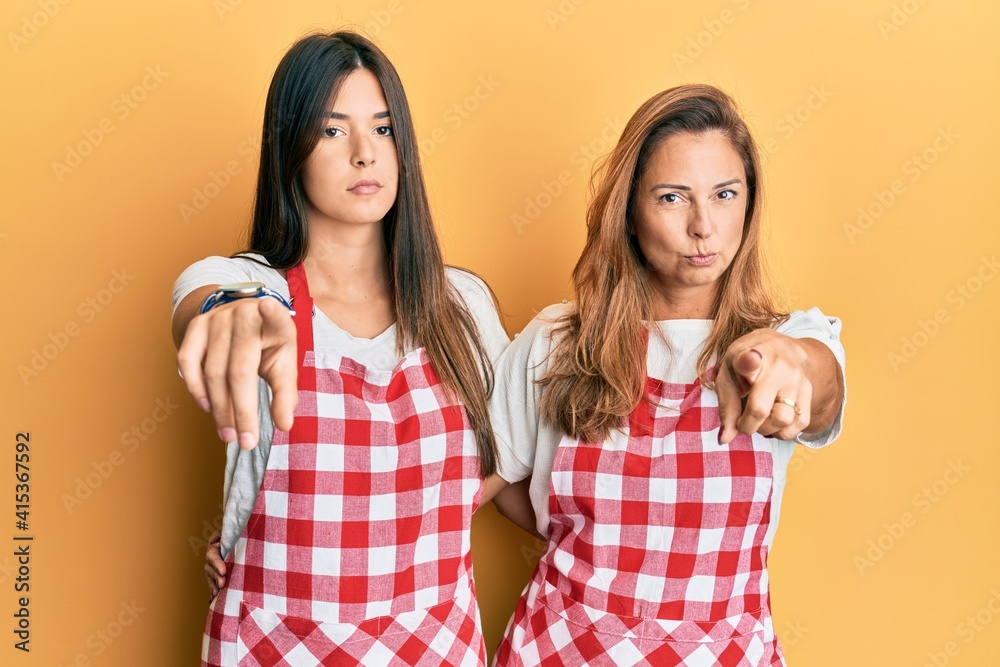 Hispanic family of mother and daughter wearing baker uniform over yellow background pointing with finger to the camera and to you, confident gesture looking serious Hispanic family of mother and daughter wearing baker uniform over yellow background pointing with finger to the camera and to you, confident gesture looking serious