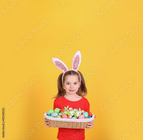 Adorable little girl with bunny ears holding wicker basket full of Easter eggs on orange background