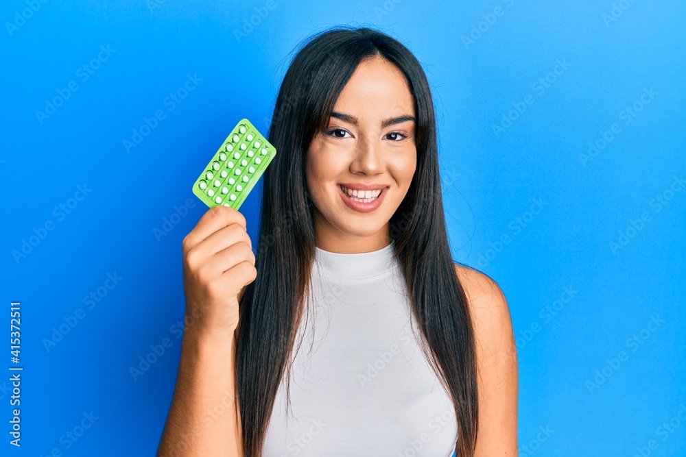 Young beautiful hispanic girl holding birth control pills looking positive and happy standing and smiling with a confident smile showing teeth Young beautiful hispanic girl holding birth control pills looking positive and happy standing and smiling with a confident smile showing teeth