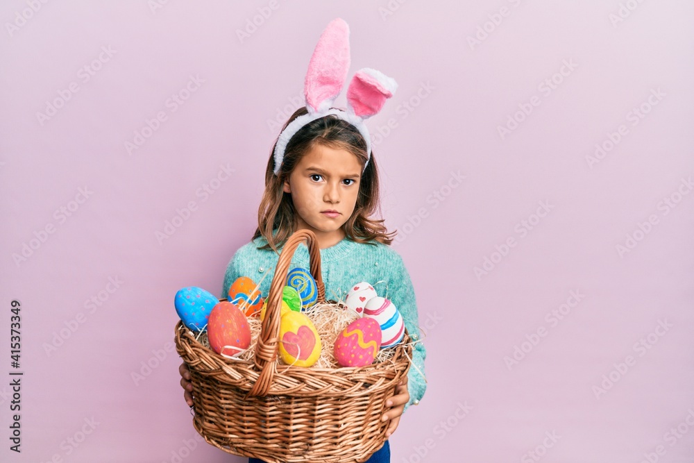 Little beautiful girl wearing cute easter bunny ears holding wicker basket with colored eggs skeptic and nervous, frowning upset because of problem. negative person.