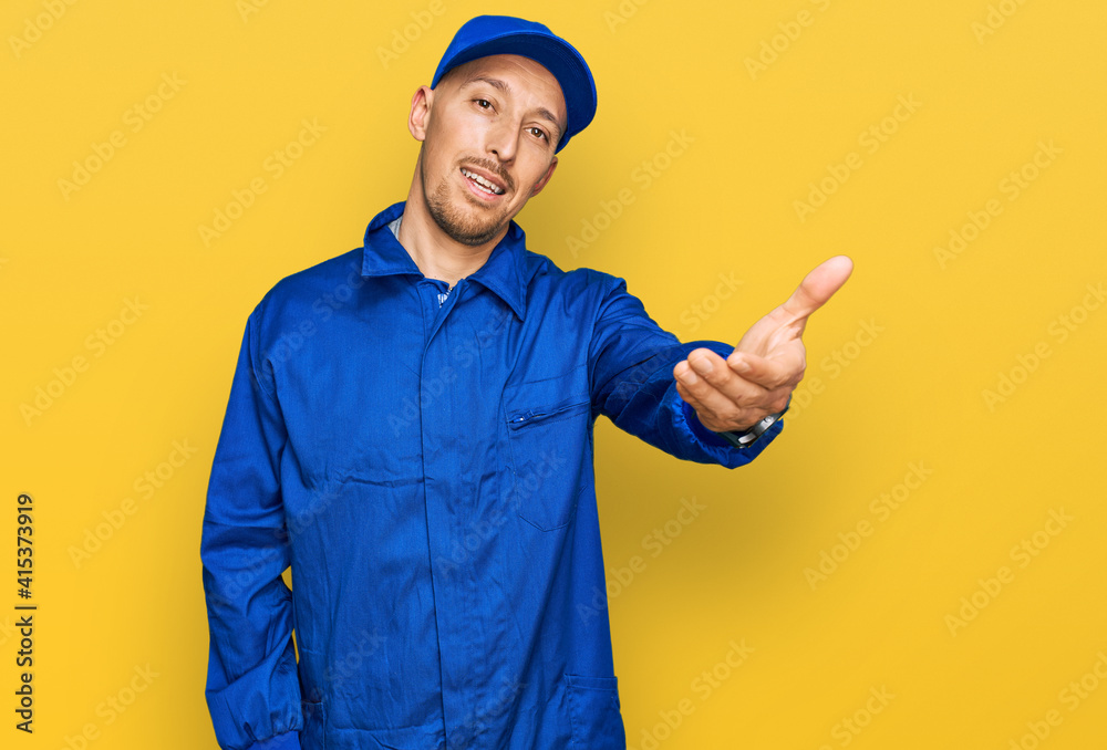 Bald man with beard wearing builder jumpsuit uniform smiling friendly offering handshake as greeting and welcoming. successful business.