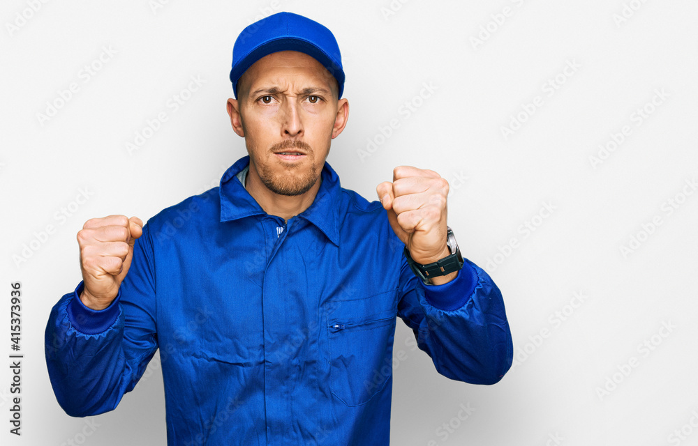 Bald man with beard wearing builder jumpsuit uniform angry and mad raising fists frustrated and furious while shouting with anger. rage and aggressive concept.