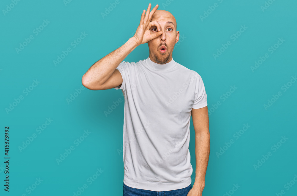 Bald man with beard wearing casual white t shirt doing ok gesture shocked with surprised face, eye looking through fingers. unbelieving expression. Bald man with beard wearing casual white t shirt doing ok gesture shocked with surprised face, eye looking through fingers. unbelieving expression.