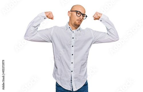 Bald man with beard wearing business shirt and glasses showing arms muscles smiling proud. fitness concept.