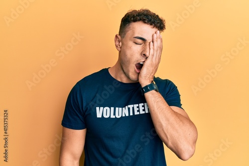 Hispanic young man wearing volunteer t shirt yawning tired covering half face, eye and mouth with hand. face hurts in pain.