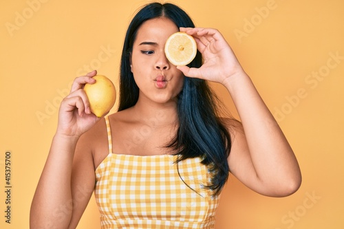 Young beautiful asian girl holding lemon making fish face with mouth and squinting eyes  crazy and comical.