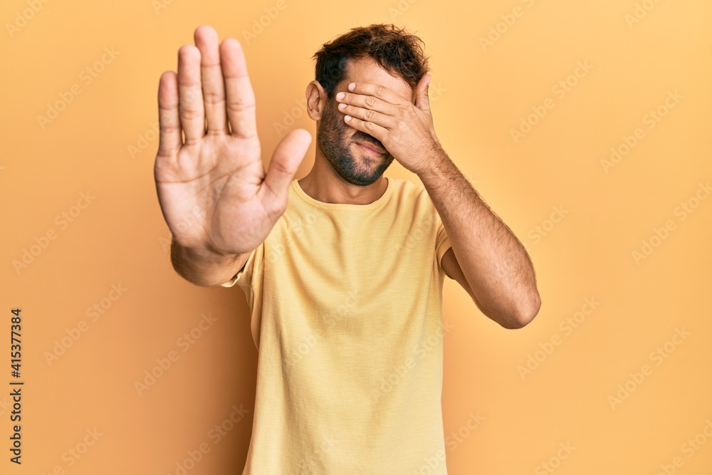 Fototapeta premium Handsome man with beard wearing casual yellow tshirt over yellow background covering eyes with hands and doing stop gesture with sad and fear expression. embarrassed and negative concept.