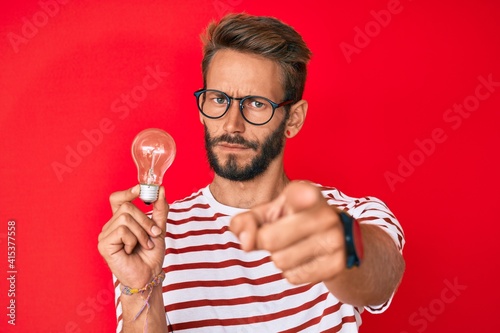 Handsome caucasian man with beard holding lightbulb for inspiration and idea pointing with finger to the camera and to you, confident gesture looking serious