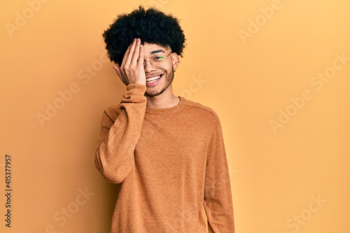 Young african american man with afro hair wearing casual winter sweater covering one eye with hand, confident smile on face and surprise emotion. © Krakenimages.com