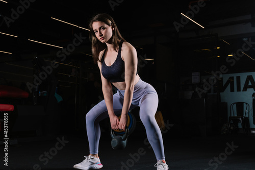 Young woman with athletic body poses sexy with kettlebell. Young woman looks beautiful in the gym. Sporty woman looking at camera.