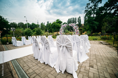 decoration of an outdoor ceremony in a green garden, an arch of fresh flowers, a white path for newlyweds, chairs in white covers