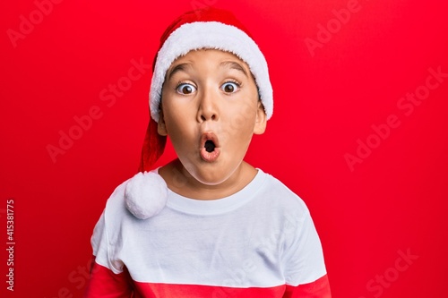 Little boy hispanic kid wearing christmas hat scared and amazed with open mouth for surprise, disbelief face photo