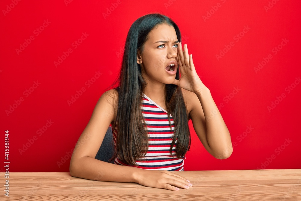 Beautiful hispanic woman wearing casual clothes sitting on the table shouting and screaming loud to side with hand on mouth. communication concept. Beautiful hispanic woman wearing casual clothes sitting on the table shouting and screaming loud to side with hand on mouth. communication concept.