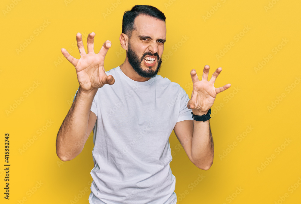 Young man with beard wearing casual white t shirt smiling funny doing claw gesture as cat, aggressive and sexy expression Young man with beard wearing casual white t shirt smiling funny doing claw gesture as cat, aggressive and sexy expression