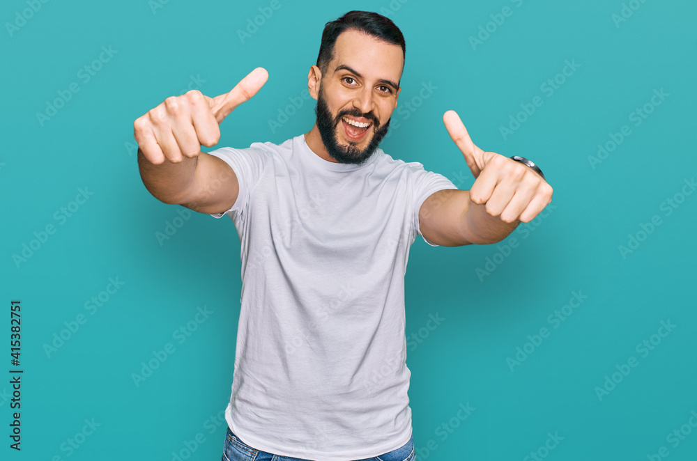 Young man with beard wearing casual white t shirt approving doing positive gesture with hand, thumbs up smiling and happy for success. winner gesture. Young man with beard wearing casual white t shirt approving doing positive gesture with hand, thumbs up smiling and happy for success. winner gesture.