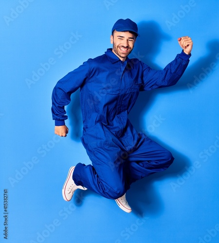 Young handsome hispanic man wearing painter uniform and cap smiling happy. Jumping with smile on face doig winner sign with fists up over isolated blue background photo